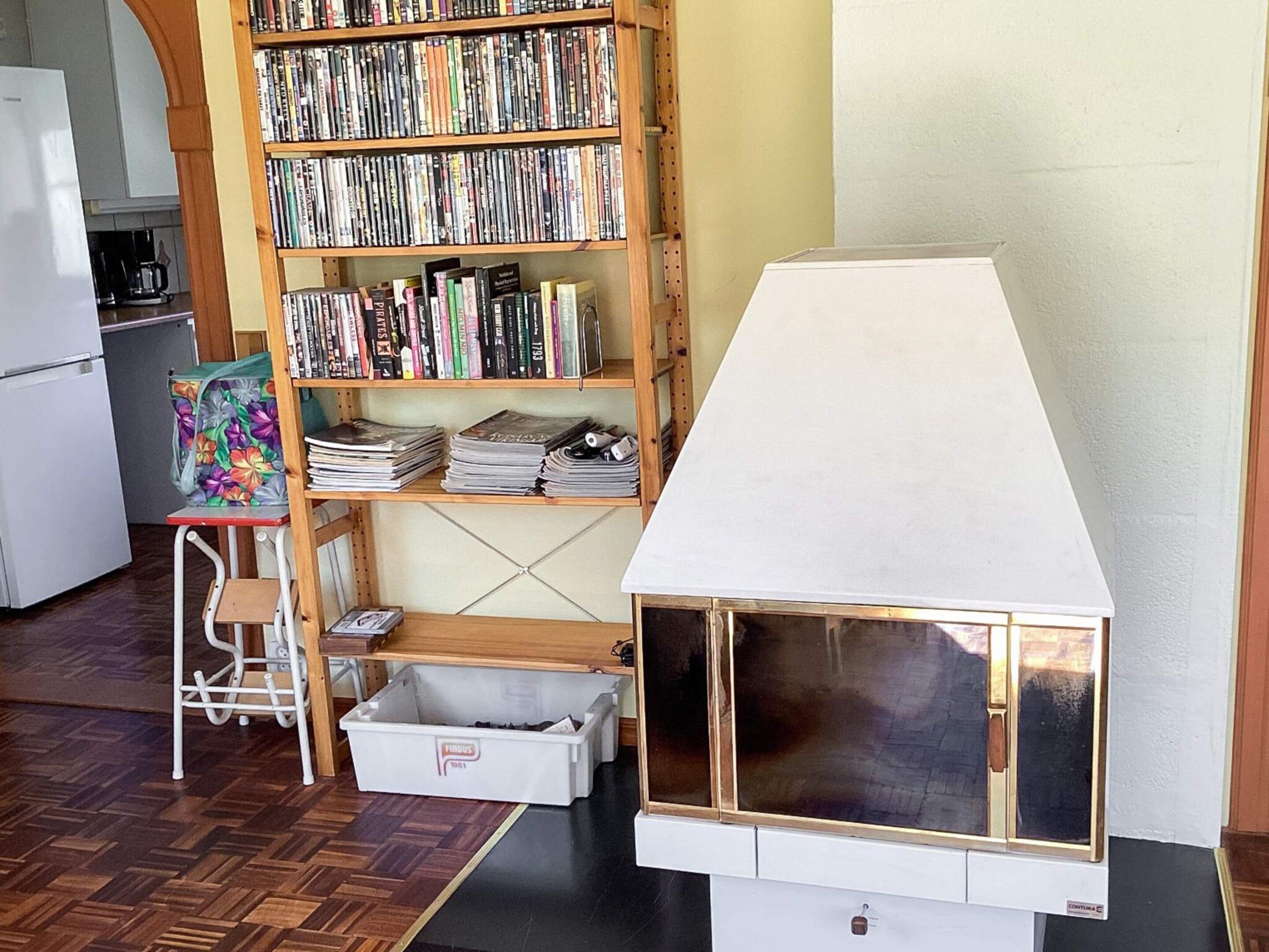 Quarto com estante de madeira repleta de livros e revistas, uma pequena mesa com uma bolsa florida, um armário branco e uma casa modelo com janela.