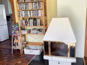 A room with a wooden bookshelf filled with books and magazines, a small table with a floral bag, a white cabinet, and a model house with a window. - 6 Person Holiday Home in Ljungbyhed (Ljungbyhed)