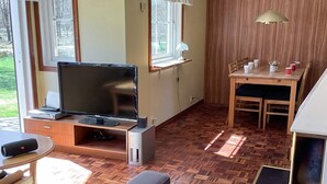 A living room with a flat-screen TV, wooden flooring, a dining table with chairs, and a window with a view of trees. - 6 Person Holiday Home in Ljungbyhed (Ljungbyhed)