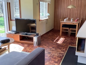 A living room with a flat-screen TV, wooden flooring, a dining table with chairs, and a window with a view of greenery. - 6 Person Holiday Home in Ljungbyhed (Ljungbyhed)