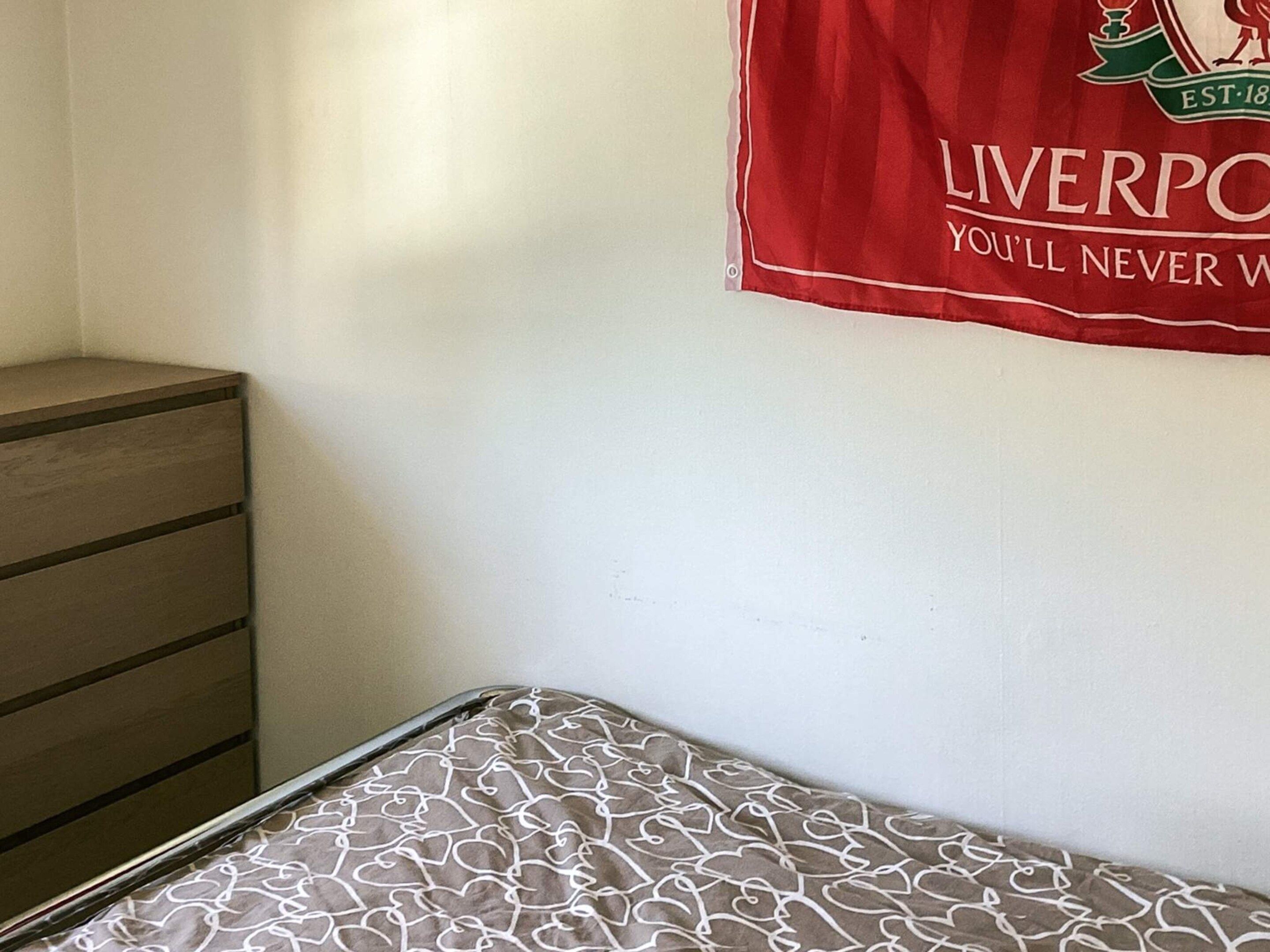 Ein Zimmer mit einem Bett, einem Holzschrank und einer Wand mit einem roten Banner des Fußballvereins Liverpool.