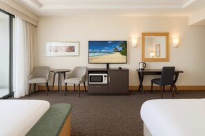Ein Hotelzimmer mit einem Bett, zwei Sesseln, einem Schreibtisch mit Computer, einem Fernseher und einem großen Fenster mit Vorhängen.