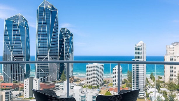 A balcony with two chairs overlooking a cityscape with modern buildings and a view of the ocean. - Stunning Ocean View Studio Pool, Spa & Wi-Fi (Surfers Paradise)