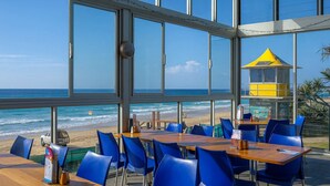 A beachside restaurant with large windows, blue chairs, wooden tables, and a lifeguard tower in the background. - Magic Nobby Beach 3BR Apt (Miami)