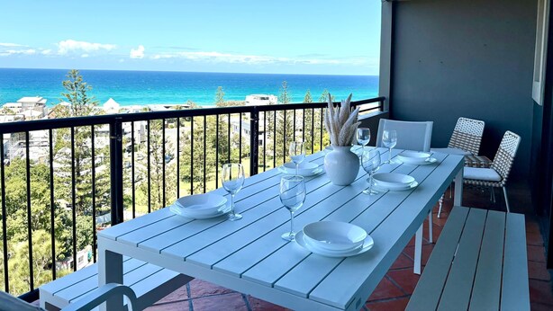 A balcony table set with white plates and glasses, overlooking a coastal view with buildings and trees. - Magic Nobby Beach 3BR Apt (Miami)