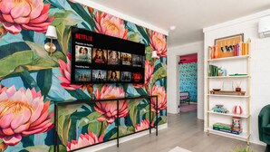 A living room with a large floral wallpaper, a Netflix display, a bookshelf, and a green armchair. - NEW Owner Special King Bed Colorful Quay Themed Duplex Workspace Fuquay-Varina (Fuquay-Varina)