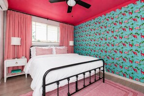 A bedroom with a bed, pink curtains, a ceiling fan, a window with blinds, and a wall with a tropical wallpaper.