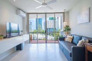 A modern living room with a flat-screen TV, a sofa, a ceiling fan, and a balcony with a city view. - 1BR Tropical Escape w/ Pools (Surfers Paradise)