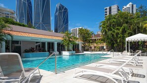 Et poolområde med liggestole, en pool og moderne bygninger i baggrunden.