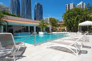 A pool area with lounge chairs, a pool, and modern buildings in the background. - 1BR Tropical Escape w/ Pools (Surfers Paradise)