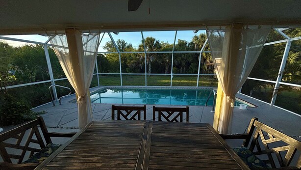 Área de piscina externa com mesa e cadeiras de madeira, cercada por uma estrutura de vidro com cortinas.