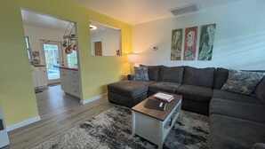 Uma sala de estar com um sofá cinza, uma mesa de centro e um tapete. Dá para ver uma cozinha ao fundo, com armários brancos e uma janela.