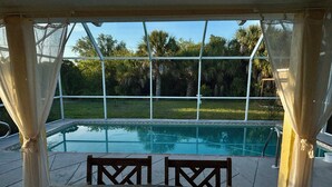 Área de piscina externa com vista para um gramado e árvores, um deck de madeira com cadeiras e uma área de estar coberta.