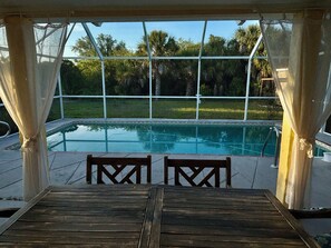 Ein Poolbereich im Freien mit Blick auf eine Rasenfläche und Bäume, eine Holzterrasse mit Stühlen und ein überdachter Sitzbereich.