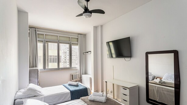 A hotel room with two beds, a TV, a large mirror, a ceiling fan, and a window with curtains. - Copacabana 2 quartos e vista mar (Rio de Janeiro)