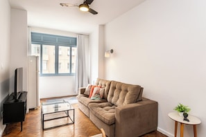 Ein Wohnzimmer mit einer Couch, einem Couchtisch, einem Fernseher, einem Deckenventilator und einem Fenster mit Vorhängen.