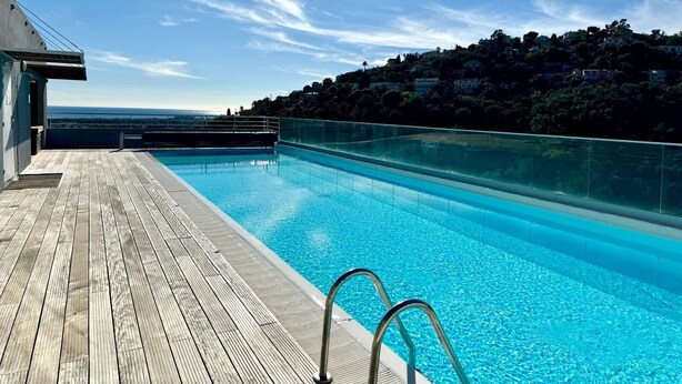 A swimming pool with a wooden deck, metal railings, and a view of a hillside with buildings. - Elegant new T2 with sea view, pool and gym in a refined setting. (Mandelieu-la-Napoule)