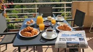 An outdoor dining table with breakfast items, a newspaper, and a scenic city view. - Elegant new T2 with sea view, pool and gym in a refined setting. (Mandelieu-la-Napoule)