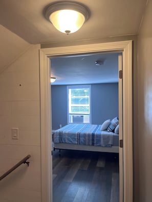 Un pasillo estrecho que conduce a un dormitorio con una cama, una ventana con persianas y una lámpara en el techo.