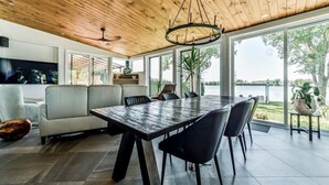 A modern dining area with a large table and chairs, a ceiling fan, and a view of a body of water through large windows. - Ô Rivensia | Bord de l`eau | Piscine | SPA (La Vallée-du-Richelieu Regional County Municipality)