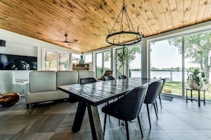 A modern dining area with a large table and chairs, a ceiling fan, and a view of a body of water through large windows.