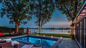 A swimming pool with lounge chairs, a pergola, and a view of a lake at dusk. - Ô Rivensia | Bord de l`eau | Piscine | SPA (La Vallée-du-Richelieu Regional County Municipality)