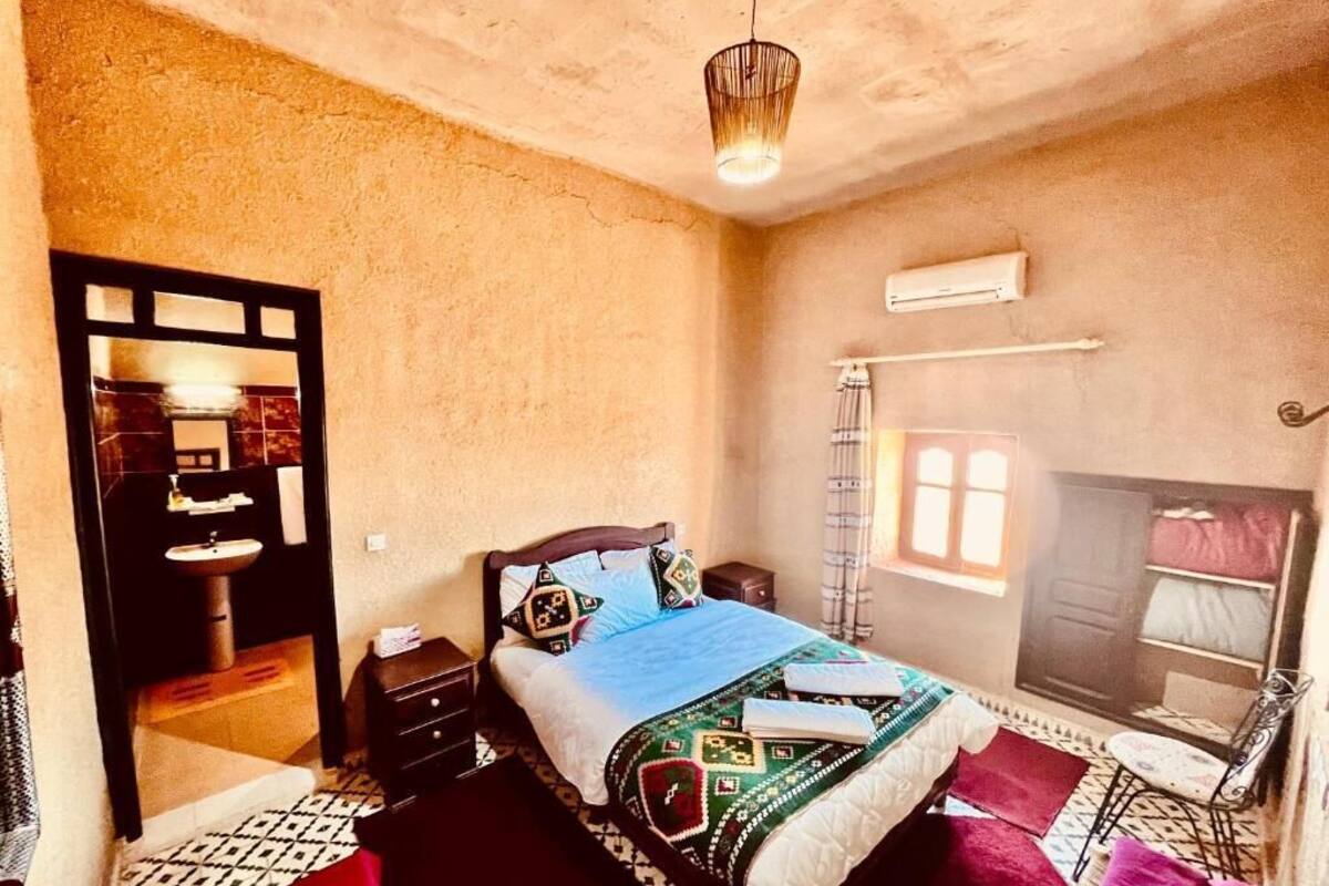 A bedroom with a bed, bedside tables, a hanging light fixture, an air conditioning unit, and a window with curtains.