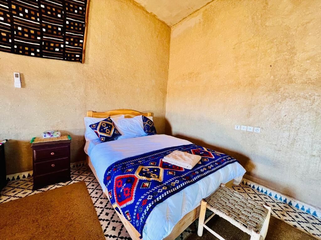 Un dormitorio con cama, mesita de noche, silla de mimbre y pared decorada.