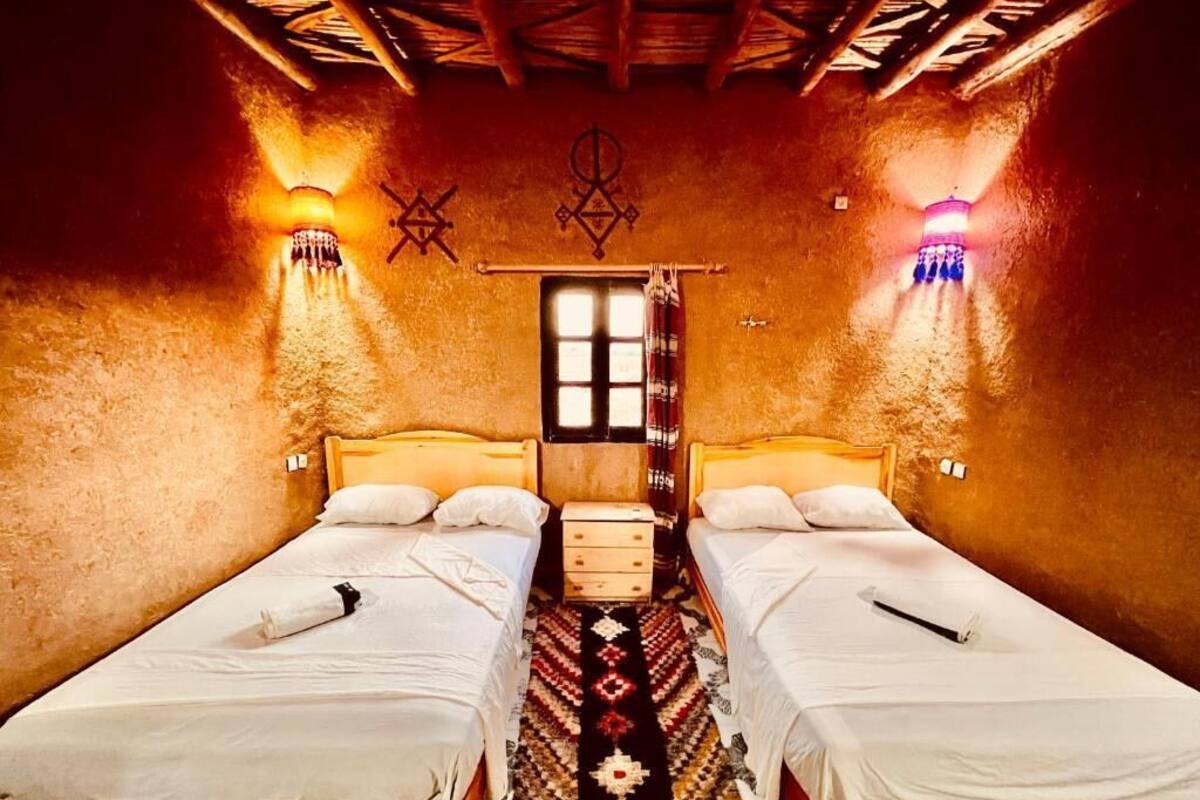A room with two single beds, a wooden ceiling, a window, and wall-mounted lights.