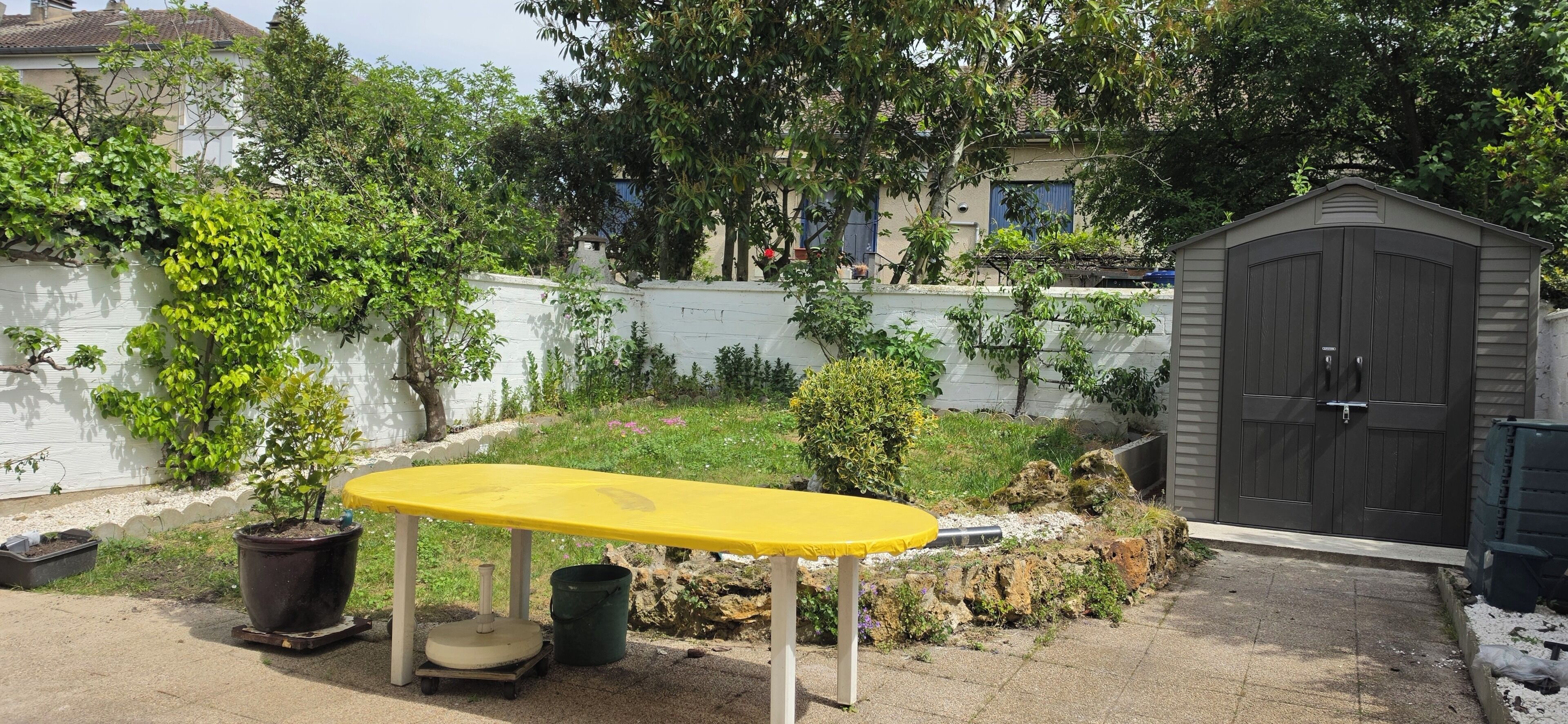 A yellow table with a potted plant and a wheelbarrow in a garden area with a white wall and a gray shed in the background.