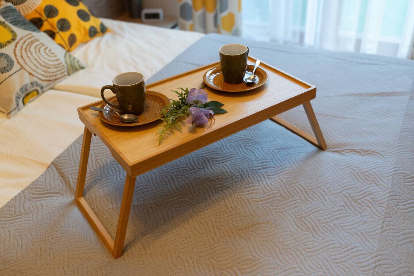 Un plateau en bois avec deux tasses et une cuillère, posé sur un lit recouvert d’une literie à motifs.