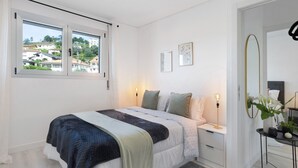 A bedroom with a bed, bedside tables, a desk, a mirror, and a window with a view of houses.