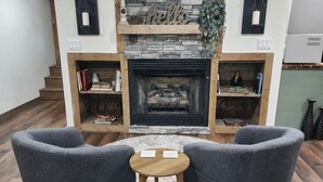 Una sala de estar con chimenea, dos sillones, una mesita redonda y una estantería.