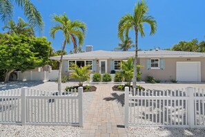 A single-story house with a paved driveway, white picket fence, and palm trees. - Sandcastle By The Sea (Holmes Beach)