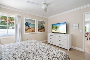 A bedroom with a bed, a TV on a white dresser, and beach-themed artwork on the wall. - Sandcastle By The Sea (Holmes Beach)