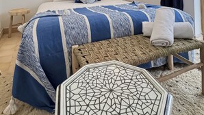 Un dormitorio con una cama, dos luces empotradas en la pared, una lámpara colgante, una ventana con cortinas y un taburete pequeño.