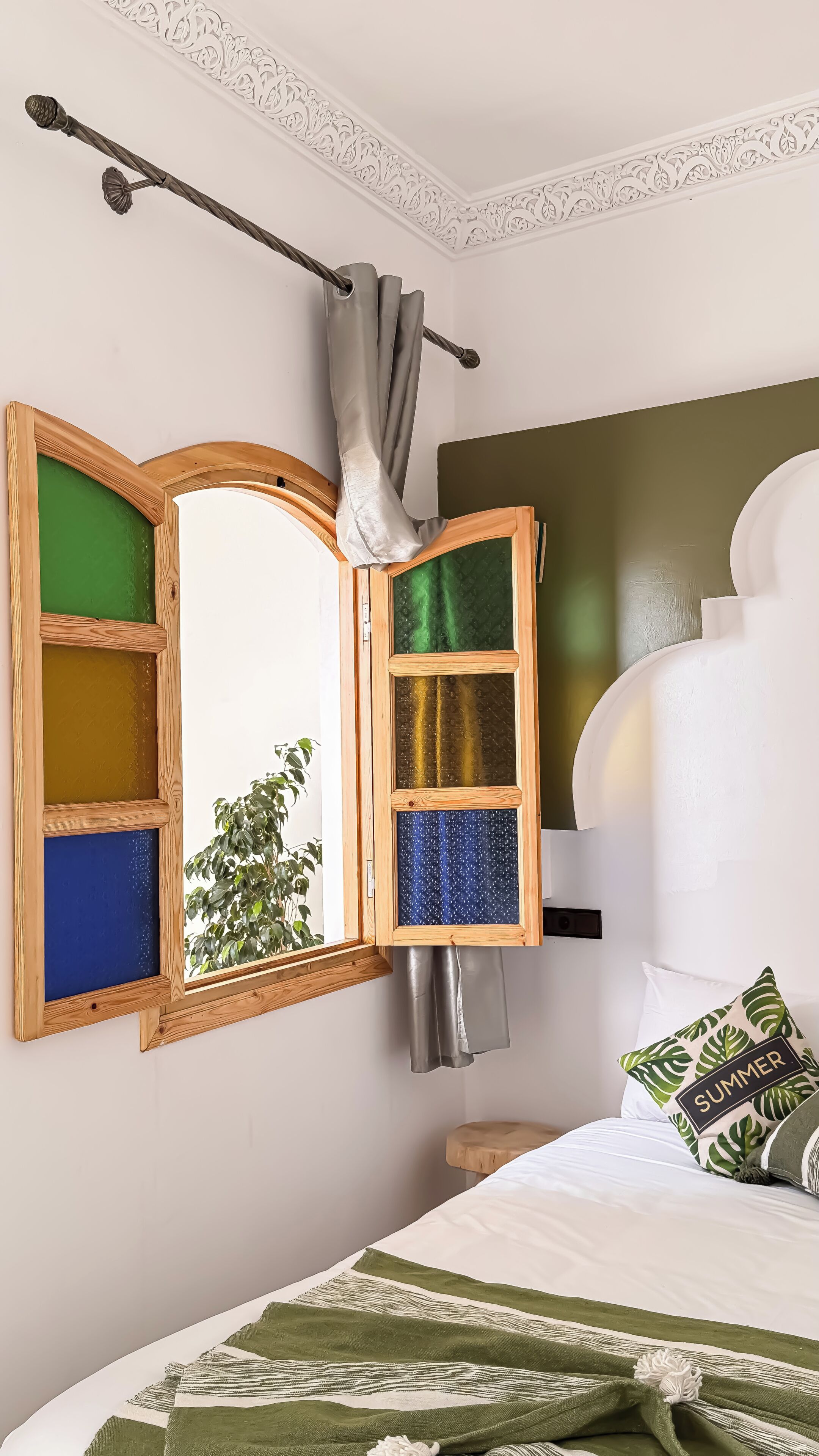 Una habitación con cama, ventana con cortinas, espejo de madera y almohada estampada con hojas.