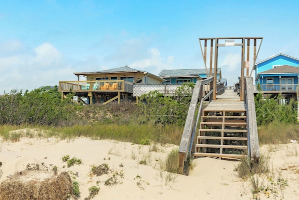 Ein Strandhaus mit einer Holzterrasse, die zu einer Veranda führt, umgeben von Sand und Vegetation.