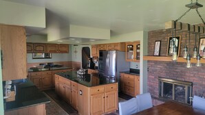 A modern kitchen with wooden cabinets, a large island, stainless steel appliances, and a brick fireplace. - Stunning 5-bedroom home on 18 acres with complete privacy, seclusion and nature (Burlington)