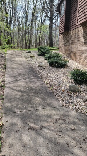 A concrete pathway leading to a brick building, surrounded by trees and small bushes.