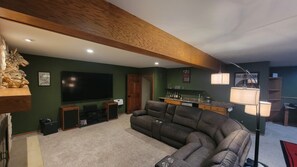 Una sala de estar con un televisor de pantalla plana grande, una pared verde oscuro, una viga de techo de madera, una zona de bar con mostrador y un sofá.