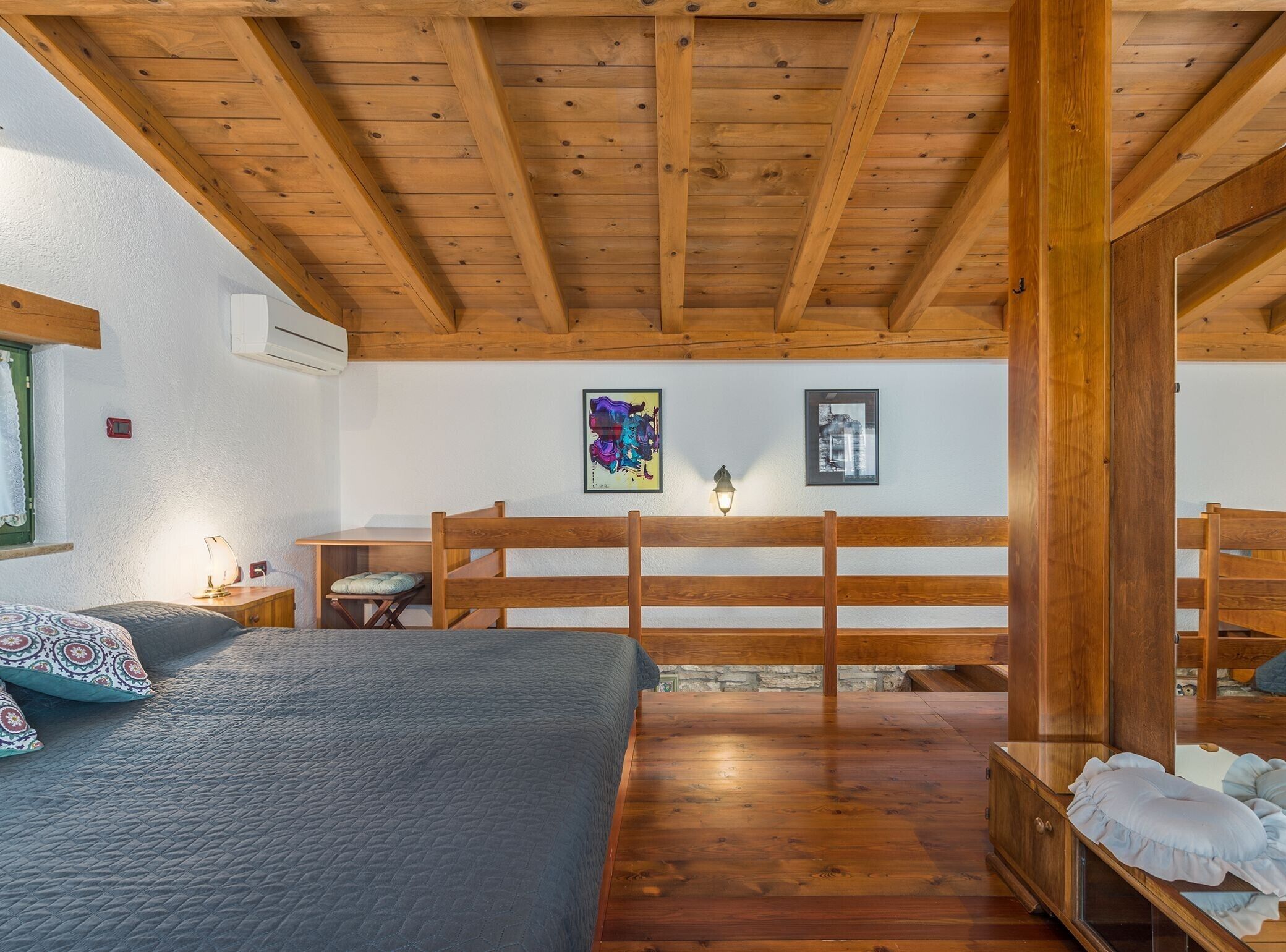 A bedroom with a wooden ceiling, a bed with patterned pillows, a nightstand, and a wall-mounted lamp.