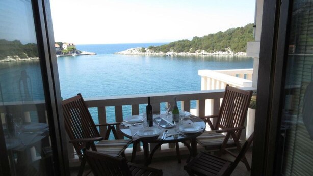 Un balcon avec une table dressée pour le dîner, offrant une vue sur une étendue d’eau et un paysage vallonné.