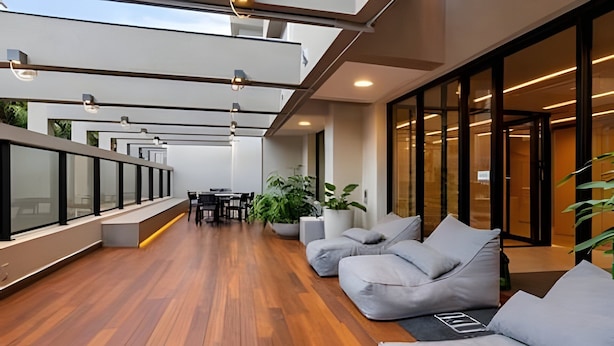 Espaço interior moderno com piso de madeira, área de estar com almofadas, plantas em vasos e guarda-corpo de vidro.