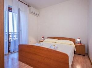 Ein Schlafzimmer mit einem hölzernen Bett, weißer Bettwäsche, zwei Nachttischlampen, einer Klimaanlage und Blick durch ein Fenster mit transparenten Vorhängen.