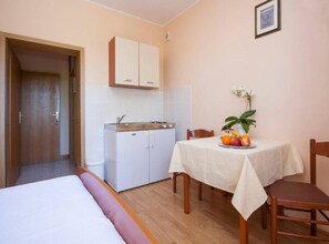 Habitación de hotel compacta con cama, kitchenette pequeña, mesa de comedor con fruta y una sola silla.