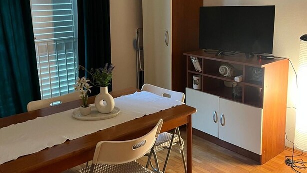 Um quarto com mesa de jantar de madeira, televisão sobre um móvel de madeira, uma cadeira, janela com persianas e um quadro na parede.