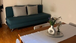 Um sofá azul-esverdeado com duas almofadas, uma mesa branca com um vaso de flores e uma parede com um quadro emoldurado.