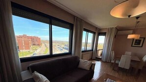 Hotellihuone, jossa on suuri ikkuna, sohva, ruokapöytä ja lamppu.