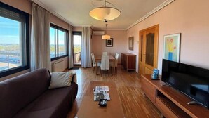 Hotellihuone, jossa on sohva, sohvapöytä, televisio, ruokailutila ja suuri ikkuna, jossa on verhot.